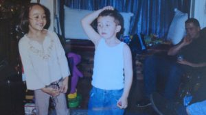 A young girl and boy stand indoors; the girl smiles and looks at the boy, who wears a white tank top and blue jeans, scratching his head. A man sits in the background on the right side of the image.