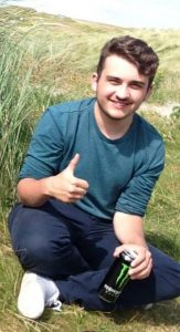 A young man sits on grass outdoors, smiling, giving a thumbs-up with one hand, and holding a can of Monster Energy drink in the other. Tall grass and greenery surround him on a sunny day.