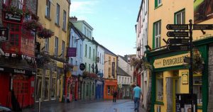 A colorful, pedestrian street lined with vibrant shops and pubs in Galway, Ireland; people stroll along the cobblestone path under a clear sky, with hanging flower baskets and signs visible.