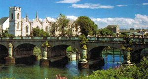 A stone bridge with multiple arches spans a calm river, with a row of white historic buildings and a church with pointed arches and spires in the background, surrounded by greenery under a blue sky.