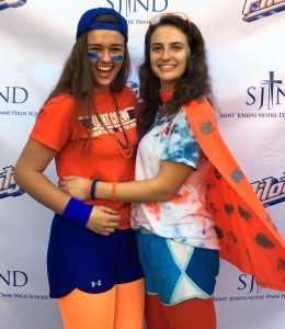 Two young women smiling and hugging, dressed in bright, sporty outfits with face paint, beads, and an orange cape. They stand in front of a backdrop that reads Saint Joseph Notre Dame High School.