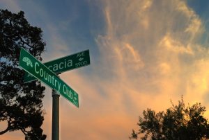 Street signs for Acacia Ave and Country Club Dr stand at a corner, silhouetted against a colorful sunset sky with wispy clouds and trees in the background.