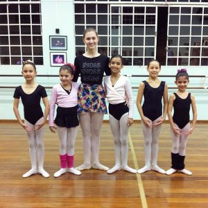 Six girls in ballet attire stand in a dance studio with wooden floors and large windows, posing in a line and smiling at the camera. The girl in the center is taller and wears a colorful skirt.