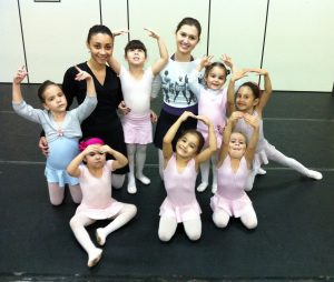 A group of young girls in ballet outfits pose with two adult instructors on a dance studio floor, smiling and holding ballet arm positions.
