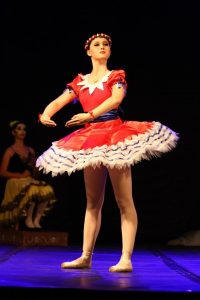 A ballerina in a red, white, and blue tutu and floral headband poses on stage with arms curved. Another dancer in a yellow costume sits in the background, both under stage lighting against a dark backdrop.