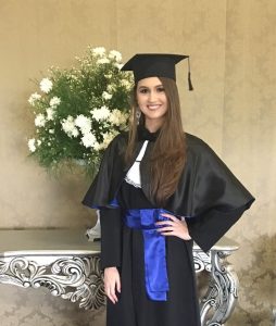 A woman wearing a black graduation gown and cap with a blue sash stands smiling beside a decorative table with a bouquet of white flowers.