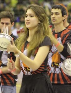 A young woman playing a percussion instrument in a group, wearing a black and red shirt with a design. Other musicians are visible in the background, also holding percussion instruments. The setting appears to be indoors.