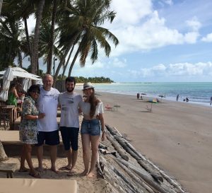 Four people stand together smiling on a sandy beach under palm trees, with the ocean and scattered beachgoers in the background on a sunny day.
