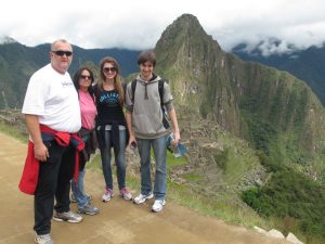 Four people stand on a dirt path at Machu Picchu, Peru, with lush green mountains and ancient stone ruins in the background. They are smiling and dressed in casual clothing.