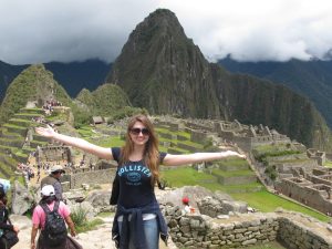 A woman with outstretched arms smiles in front of the ancient Incan ruins of Machu Picchu, surrounded by green mountains under a cloudy sky. Tourists are visible exploring the site in the background.