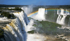 Powerful waterfalls cascade over rocky cliffs surrounded by lush green vegetation, with mist rising and a vibrant rainbow arching across the scene under a clear blue sky.