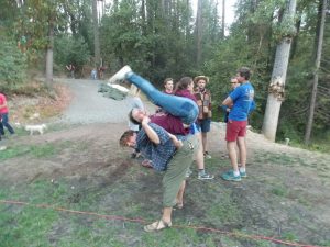 A group of young people stand outdoors among trees; in the foreground, one person is playfully flipping another person upside down over their back. Others watch and smile.