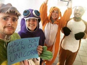 Four people in animal onesies smile at the camera. One holds a blue sign that says “HAVE YOU SEEN OUR UNICORN?” Costumes include a frog, a purple creature, an orange animal, and a brown-and-white animal.