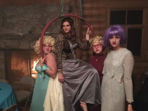 Four people in colorful wigs and vintage-style dresses pose indoors; one sits on a hoop, the others stand beside her. There’s a stone fireplace and wooden walls in the cozy background.