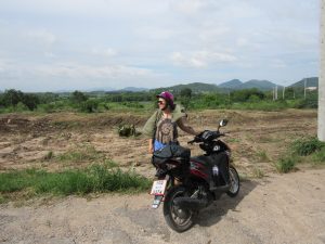 A person wearing a purple helmet and sunglasses stands next to a parked black and red scooter on a dirt road, with green fields, trees, and mountains in the background under a partly cloudy sky.