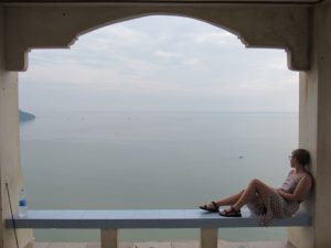 A woman in a dress sits on a tiled ledge, relaxing and gazing at a calm sea through a large arched window. The sky is overcast, and a lone boat is visible on the water in the distance.