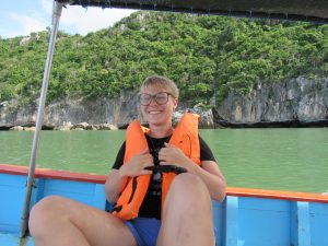 A person with short hair and glasses, smiling and wearing an orange life jacket, sits on a blue boat with green water and rocky, forested cliffs in the background.