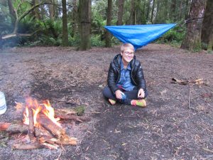 A person wearing glasses, a black jacket, and bright shoes sits on the ground near a campfire in a forest. A blue tarp is strung between trees in the background.