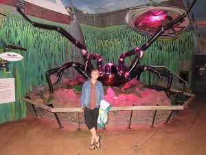 A person stands smiling in front of a giant black spider model in an indoor exhibit with grass murals and a UFO prop above. The person wears a denim jacket, black pants, and flip-flops.