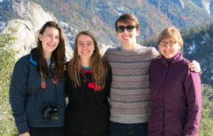 Four people stand close together outdoors with mountains in the background. They are smiling at the camera; one person wears sunglasses, another has a camera around their neck. The group wears winter clothing.