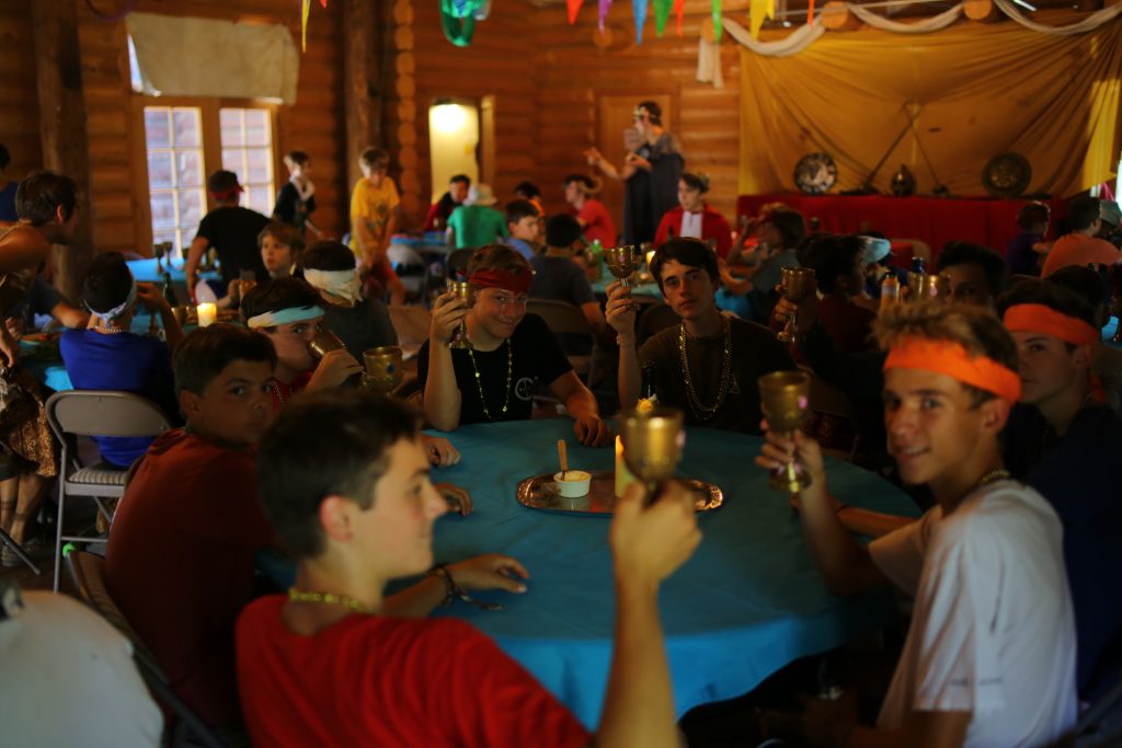 A group of teens, joined by cheerful summer camp staff, sit around a table in a festively decorated wooden hall, holding up gold-colored goblets in a toast. The room is lively, adorned with colorful streamers and a long table set at the front.