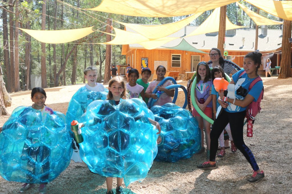 A group of children at an outdoor camp, joined by summer camp staff, some wearing large blue inflatable suits and others holding colorful inflatable toys. Trees, shade sails, and a wooden building are in the background.