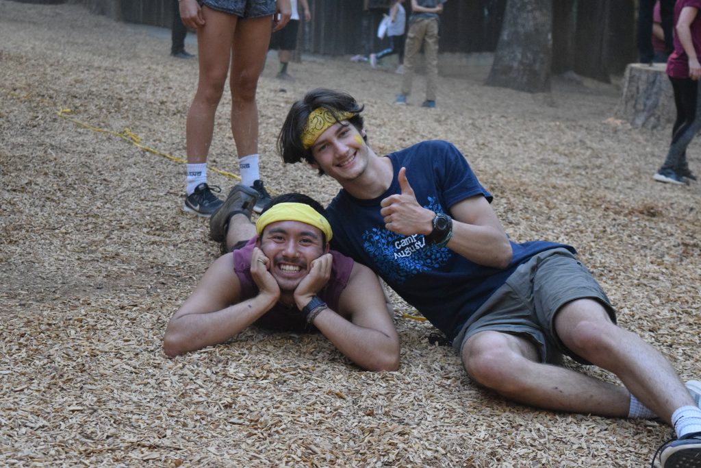Two young adults in casual clothes and yellow headbands—likely summer camp staff—are smiling on a wood chip-covered ground; one lies on their stomach with hands under chin, while the other reclines next to them giving a thumbs-up.