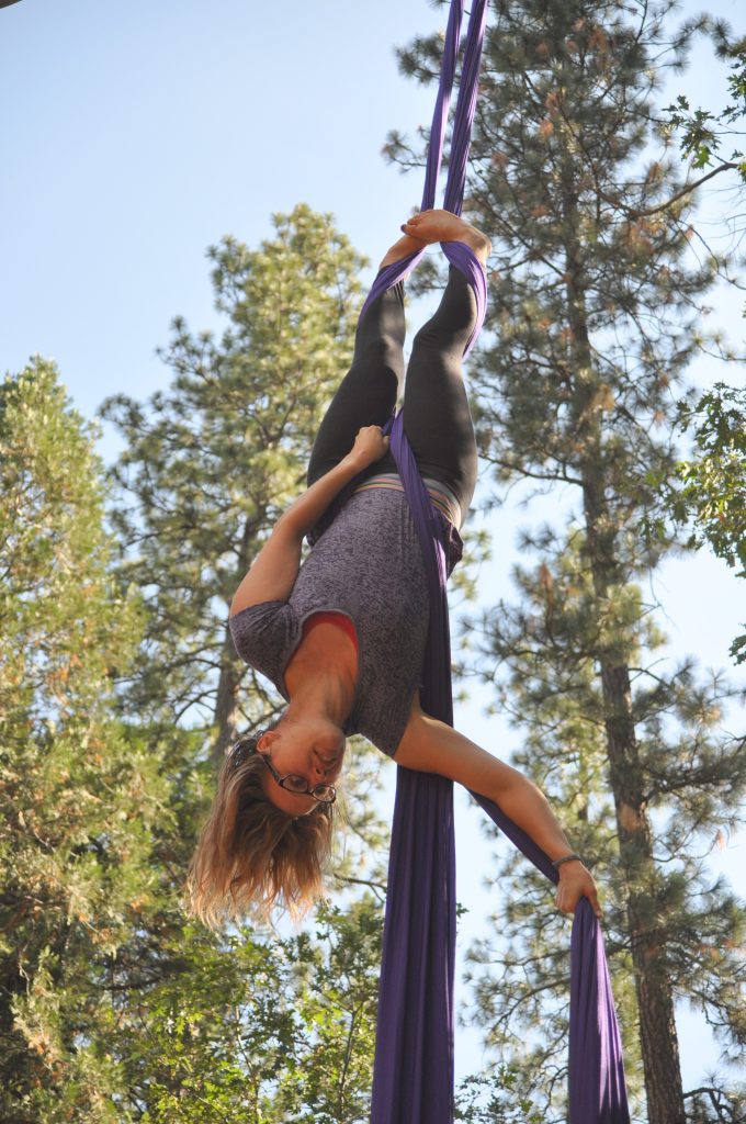 A woman performs an aerial silk routine outdoors, hanging upside down from purple fabric with tall pine trees and a clear sky in the background.