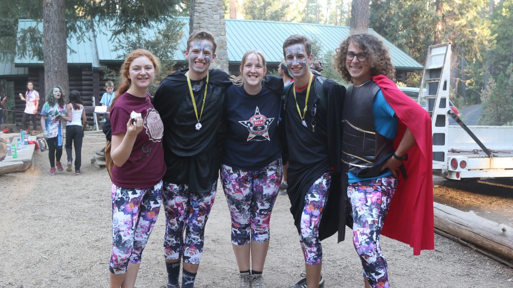 Five people in matching colorful leggings and costumes, some with face paint and medals, smile and pose together outdoors at camp, with trees, buildings, and campers in the background.