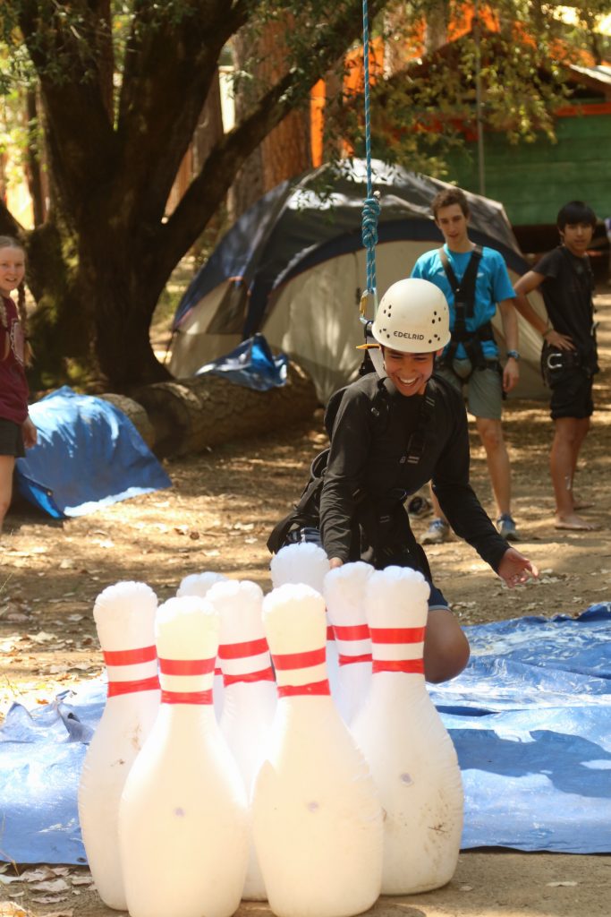A summer camp staff member wearing a helmet swings on a rope toward large inflatable bowling pins outdoors, while others watch in a wooded area with tents in the background.