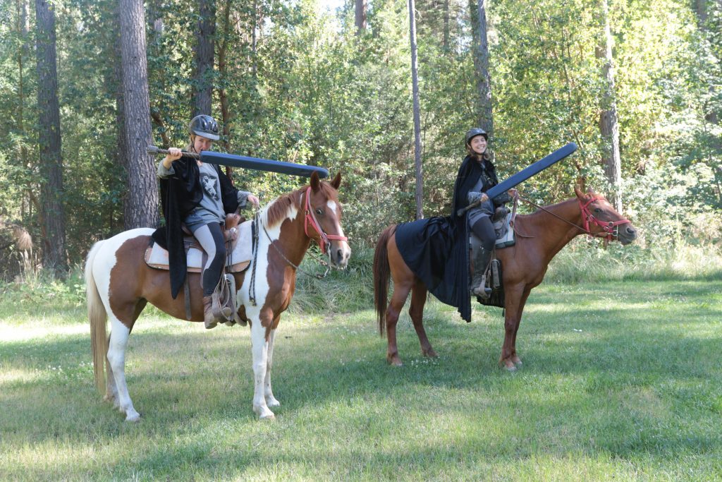 Two people in medieval-style capes and helmets sit on horses in a grassy clearing, each holding a large foam jousting lance. Tall trees fill the background.