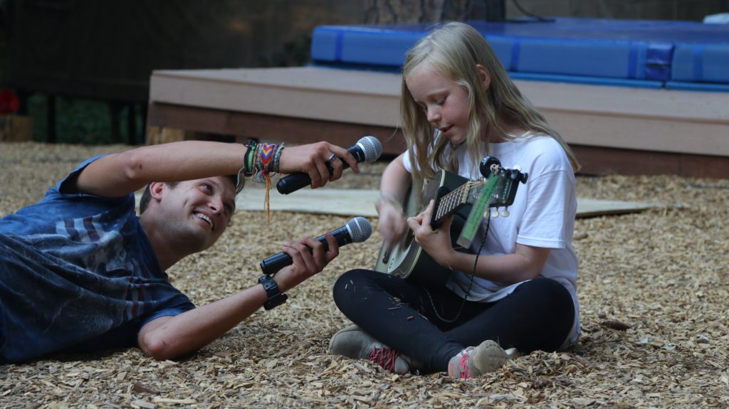 A man lying on the ground smiles while holding two microphones toward a young girl who sits on wood chips, playing an acoustic guitar and singing. They are outdoors, with a blue mat and wooden steps in the background.