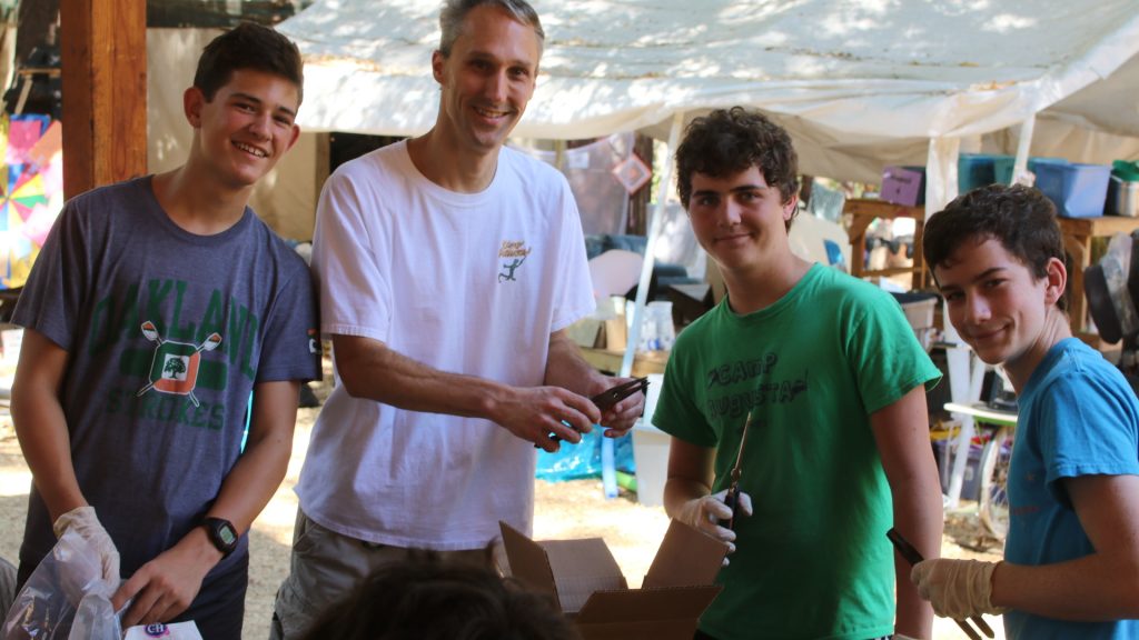 Four people, three teens and one adult, smile while working together outdoors under a canopy. They appear to be handling tools or supplies and are engaged in an activity, possibly volunteering or doing a group project.