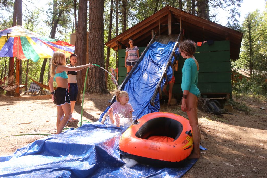 Four children play on a homemade slip-and-slide made of blue tarp outside a cabin in the woods, supervised by summer camp staff. One child sprays water with a hose as another slides toward an orange inflatable raft, while two others watch.