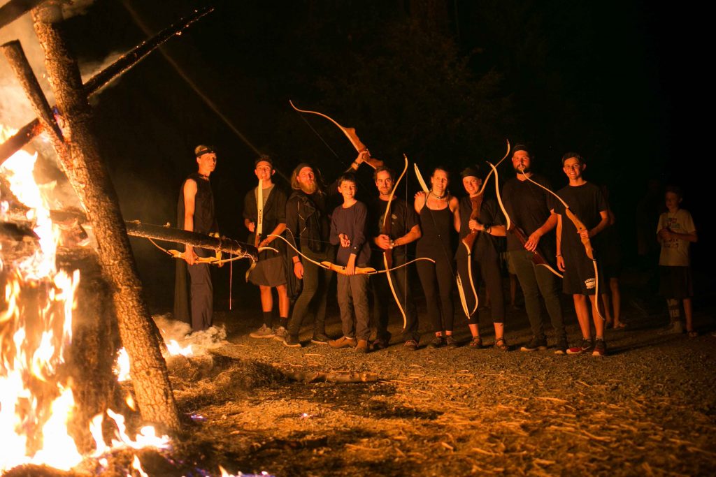 A group of people stand at night near a large, burning wooden structure, each holding a bow. The group is illuminated by the fire, with trees and darkness in the background.