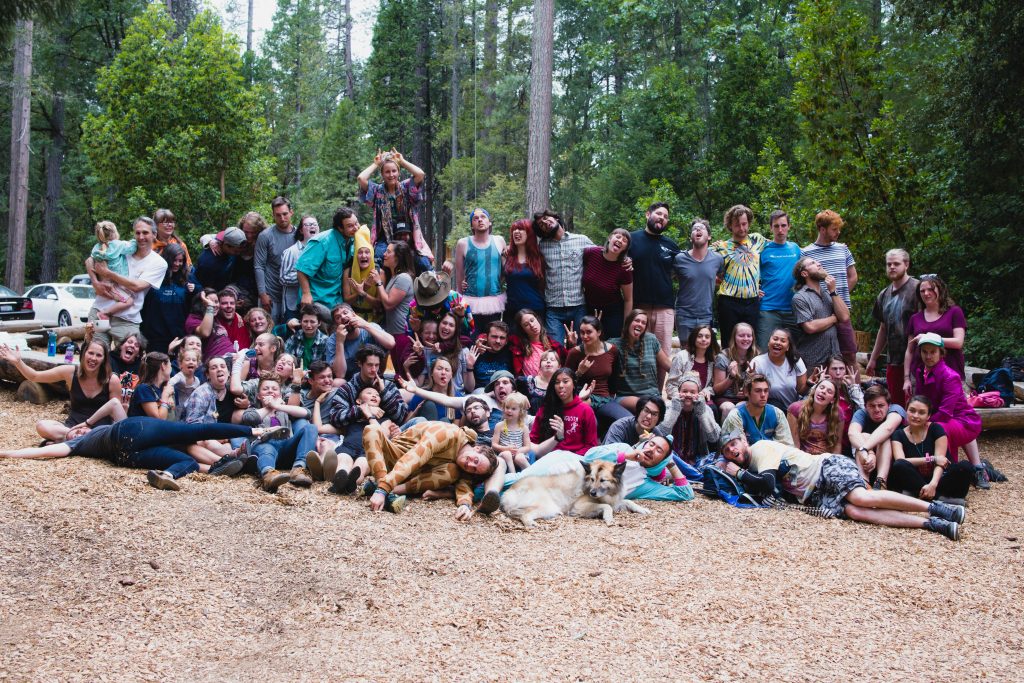A large group of people pose playfully for a photo in a wooded outdoor area, some sitting, standing, or lying on the ground, with a dog lying at the front; trees and greenery fill the background.