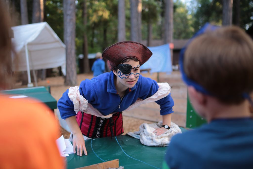 A person dressed as a pirate, with a red hat, eye patch, and blue top, leans over a table outdoors, talking to a child. Tents and trees are visible in the background, suggesting a camp or fair setting.