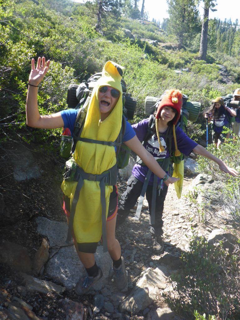 Two hikers, one dressed in a banana costume and the other in a red animal hat, smile and pose playfully on a sunny, rocky forest trail. Other hikers with backpacks are visible in the background.