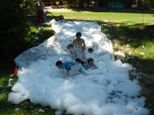 A group of children and a summer camp staff member play on a homemade slip and slide covered in white foam outdoors on a sunny day, with green grass and trees in the background.