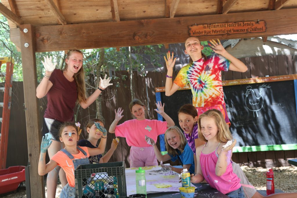 A group of smiling children and two adults stand under a wooden shelter, waving their paint-covered hands. They are gathered around a table with art supplies, with colorful artwork and a chalkboard visible in the background.