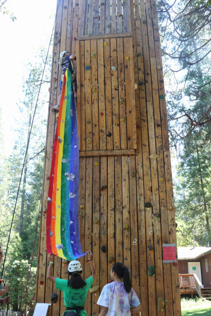 A child climbs a tall wooden rock climbing wall, while two people at the bottom hold a long, colorful rainbow flag that trails up alongside the climber. Trees and a cabin are visible in the background.