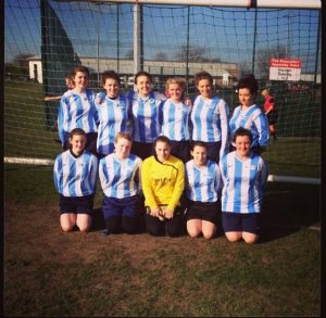 A women’s soccer team poses together in front of a goalpost on a sunny day. Most players wear blue and white striped jerseys, with one in a yellow goalie shirt, and they are standing and kneeling on grass.