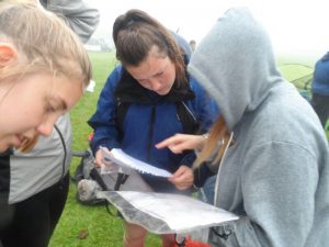 Three young people are outdoors on a foggy day, closely examining maps or documents. One person is pointing at a map while the others look intently, dressed in casual outdoor clothing. Grass and tents are visible in the background.