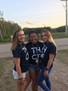 Three young women wearing matching navy blue T-shirts with white letters stand close together outdoors, smiling at the camera. Trees and a road are visible in the background.