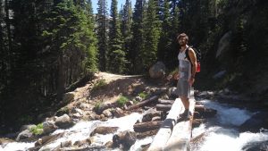 A man wearing sunglasses and a backpack stands on a log bridge over a rushing creek in a forested mountain area, with sunlight shining through the tall pine trees.
