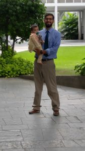 A man wearing a blue shirt, tie, and khaki pants stands on a stone walkway holding a baby dressed in light-colored clothes, with green grass and bushes in the background.