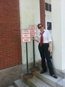 A woman in a white shirt, black pants, and red tie stands next to two identical Police Parking Only Between Signs signs placed directly beside each other, highlighting the humorous redundancy.