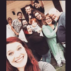 A group of eleven people, including a small child, smiling and posing closely together for a selfie on a porch. The atmosphere is joyful and casual, suggesting a family or friends gathering.