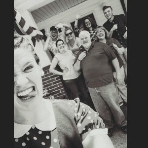 A group of nine people pose and smile enthusiastically for a selfie outside a house. The person in the front makes a funny face, while the others stand together in the background, waving and laughing. The photo is in black and white.