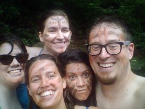 Five people with face paint smile and pose closely for a selfie outdoors; one wears glasses, one wears sunglasses, and trees or greenery are visible in the background.